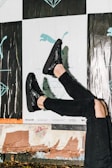 A person wearing shiny black shoes and black ripped jeans against a background featuring posters with abstract and illustrative designs, including a silhouette of a puma and geometric shapes. The scene suggests a casual urban environment.