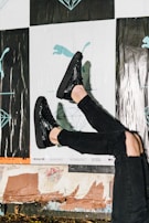 A person wearing shiny black shoes and black ripped jeans against a background featuring posters with abstract and illustrative designs, including a silhouette of a puma and geometric shapes. The scene suggests a casual urban environment.