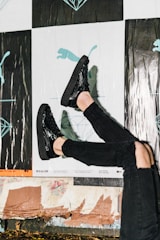 A person wearing shiny black shoes and black ripped jeans against a background featuring posters with abstract and illustrative designs, including a silhouette of a puma and geometric shapes. The scene suggests a casual urban environment.