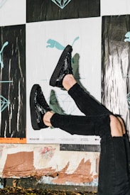 A person wearing shiny black shoes and black ripped jeans against a background featuring posters with abstract and illustrative designs, including a silhouette of a puma and geometric shapes. The scene suggests a casual urban environment.