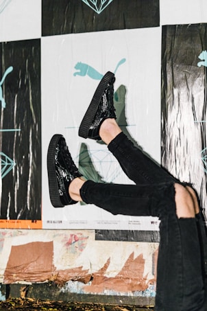 A person wearing shiny black shoes and black ripped jeans against a background featuring posters with abstract and illustrative designs, including a silhouette of a puma and geometric shapes. The scene suggests a casual urban environment.