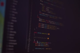 A dark background with colorful lines of code in various programming languages displayed on a screen. The text includes snippets of functions and logical statements.