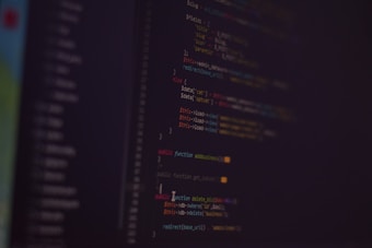 A dark background with colorful lines of code in various programming languages displayed on a screen. The text includes snippets of functions and logical statements.
