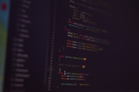 A dark background with colorful lines of code in various programming languages displayed on a screen. The text includes snippets of functions and logical statements.