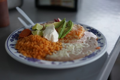 A plate of Mexican food featuring rice, refried beans topped with melted cheese, a dollop of sour cream, slices of avocado, lime wedges, and a few sliced vegetables including a carrot. Utensils are placed next to the plate on a table.