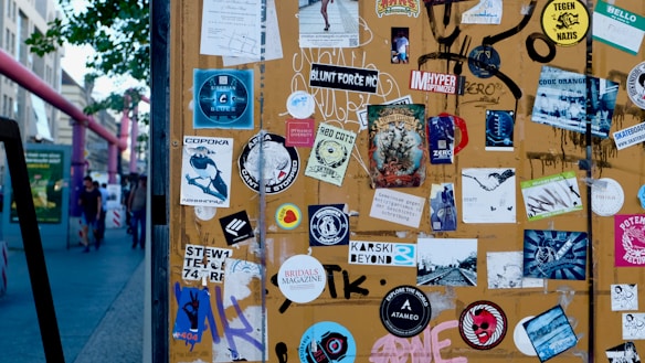 A variety of stickers cover a brown board, showcasing different designs and texts. A few stickers include illustrations, logos, and text such as 'BRIDALS MAGAZINE,' 'CODE ORANGE,' and 'STEW1 74RF.' There is a blend of colors and styles, creating a visually chaotic scene. In the background, a street scene with pedestrians and buildings can be seen.