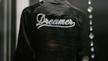 A dark jacket with the word 'Dreamer' written in white script across the back is captured in a low light setting. The texture of the fabric is slightly shiny, and there are blurred reflections of light on the right side, possibly from glass or another reflective surface.