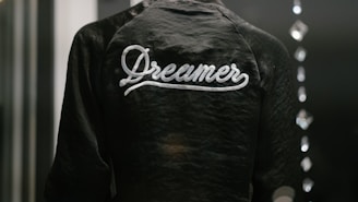 A dark jacket with the word 'Dreamer' written in white script across the back is captured in a low light setting. The texture of the fabric is slightly shiny, and there are blurred reflections of light on the right side, possibly from glass or another reflective surface.
