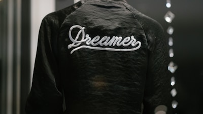 A dark jacket with the word 'Dreamer' written in white script across the back is captured in a low light setting. The texture of the fabric is slightly shiny, and there are blurred reflections of light on the right side, possibly from glass or another reflective surface.