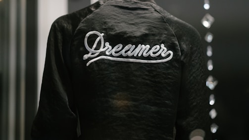 A dark jacket with the word 'Dreamer' written in white script across the back is captured in a low light setting. The texture of the fabric is slightly shiny, and there are blurred reflections of light on the right side, possibly from glass or another reflective surface.