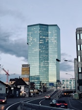 A modern skyscraper with a sleek, glass facade dominates the urban landscape. Several smaller buildings, including one with a tower of colorful container boxes, surround it. There's a red construction crane on the left. A curved road leads toward the skyscraper, with a few cars driving along it under a cloudy sky.