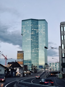 A modern skyscraper with a sleek, glass facade dominates the urban landscape. Several smaller buildings, including one with a tower of colorful container boxes, surround it. There's a red construction crane on the left. A curved road leads toward the skyscraper, with a few cars driving along it under a cloudy sky.
