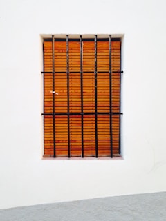 A window with vertical black bars is set in a white wall. Behind the bars, there is a wooden shutter with a horizontal slat pattern.