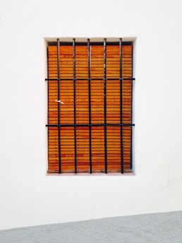 A window with vertical black bars is set in a white wall. Behind the bars, there is a wooden shutter with a horizontal slat pattern.