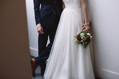 A bride in a white gown holds a bouquet while standing next to a groom in a dark suit. They are in a minimalist setting with neutral-toned walls and flooring.