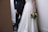 A bride in a white gown holds a bouquet while standing next to a groom in a dark suit. They are in a minimalist setting with neutral-toned walls and flooring.