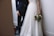 A bride in a white gown holds a bouquet while standing next to a groom in a dark suit. They are in a minimalist setting with neutral-toned walls and flooring.