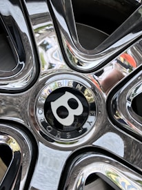 Close-up view of a shiny, reflective Bentley car wheel rim with intricate design and engraved brand name, surrounded by chrome detailing that captures reflections of the surroundings.
