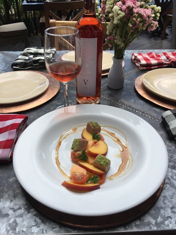 A plate with a gourmet dish of sliced peaches, cherry tomatoes, and cubes of food garnished with herbs, drizzled with a sauce. The setting includes a bottle of rosé wine, a glass partially filled with the wine, and a small bouquet of flowers in a vase. The table is set with plates and checkered napkins, creating a rustic dining atmosphere.