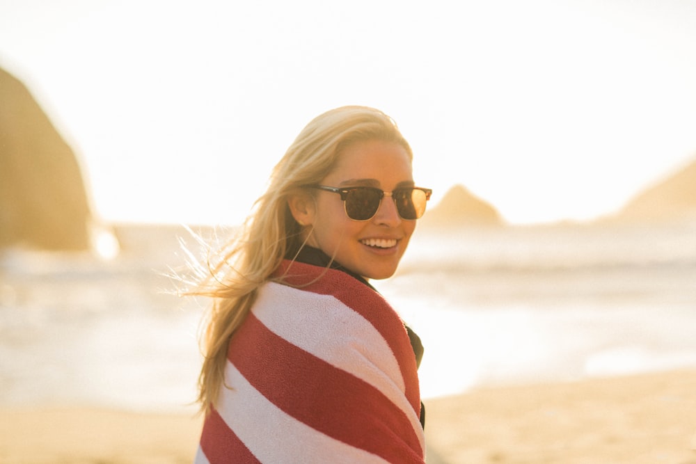woman wearing black sunglasses with red and white striped blanket cover standing near seashore during daytime