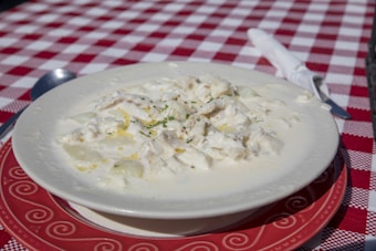 A bowl of creamy potato soup garnished with herbs is placed on a decorative red plate. The table is covered with a red and white checkered tablecloth, and there is a spoon and knife wrapped in a white napkin beside it.