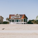 A large, two-story beachfront house with a distinctive red roof and white exterior is situated on a sandy beach. The structure features a spacious porch and a circular screened area on one side. Surrounding the house, there are palm trees and a smaller green house is partially visible on the right side.