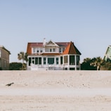 A large, two-story beachfront house with a distinctive red roof and white exterior is situated on a sandy beach. The structure features a spacious porch and a circular screened area on one side. Surrounding the house, there are palm trees and a smaller green house is partially visible on the right side.