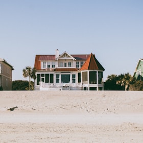 A large, two-story beachfront house with a distinctive red roof and white exterior is situated on a sandy beach. The structure features a spacious porch and a circular screened area on one side. Surrounding the house, there are palm trees and a smaller green house is partially visible on the right side.