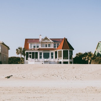 A large, two-story beachfront house with a distinctive red roof and white exterior is situated on a sandy beach. The structure features a spacious porch and a circular screened area on one side. Surrounding the house, there are palm trees and a smaller green house is partially visible on the right side.