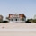 A large, two-story beachfront house with a distinctive red roof and white exterior is situated on a sandy beach. The structure features a spacious porch and a circular screened area on one side. Surrounding the house, there are palm trees and a smaller green house is partially visible on the right side.