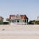 A large, two-story beachfront house with a distinctive red roof and white exterior is situated on a sandy beach. The structure features a spacious porch and a circular screened area on one side. Surrounding the house, there are palm trees and a smaller green house is partially visible on the right side.