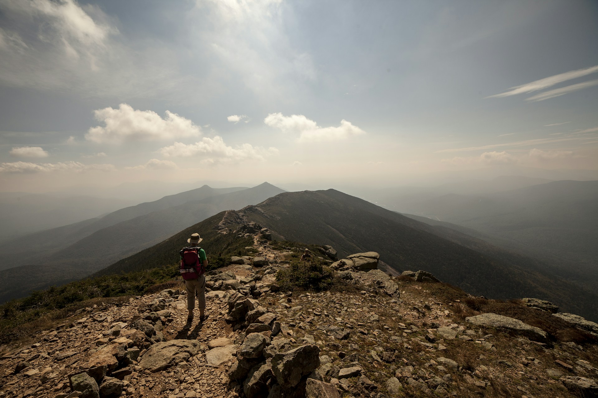 A winding mountain trail with a lone traveler pausing to take in the breathtaking panoramic view of distant peaks.