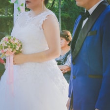 A bride in a white lace wedding dress holding a bouquet of pink and white flowers stands next to a man in a blue suit with a black bow tie. An older woman is seated in the background, looking on attentively. The setting appears to be outdoors, with greenery visible.