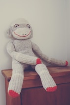 A knitted sock monkey with a cheerful expression is seated on the edge of a wooden piece of furniture. The monkey has a gray body with bright red accents on its hands, feet, and smiling mouth.