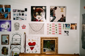 A collage of posters and artwork is arranged on a wall. The central piece features a cartoon bear, while another prominent poster shows two individuals on an album cover. Various other images include a skull sketch, a painting of Snoopy, abstract art, and several photos. Below, a decorative shelf holds small items, situated next to a gold-framed mirror.