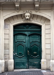 A pair of large, ornate green wooden doors with decorative panels and brass handles. Positioned above the doors is an intricately carved stone lion head as part of the archway, which is framed by detailed stonework featuring classical architectural elements.