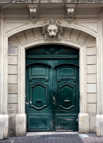 A pair of large, ornate green wooden doors with decorative panels and brass handles. Positioned above the doors is an intricately carved stone lion head as part of the archway, which is framed by detailed stonework featuring classical architectural elements.