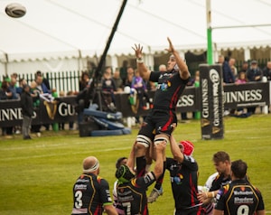 Rugby players are engaged in a line-out during a match, with one player being lifted high to catch the ball. The scene takes place on a grassy field surrounded by an audience, with branding and advertisements in the background.