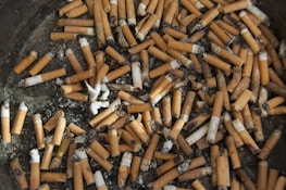Numerous cigarette butts are scattered across an ashtray, with some partially burnt and showing signs of ash residue. The majority have brown filters with white paper and blackened tips from being smoked.