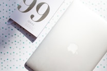 A minimalist setup features a white notebook titled '99 Ideas' placed beside a closed silver laptop with a subtle Apple logo. The surface is dotted with teal and light blue polka dots, enhancing the light and airy feel.
