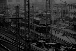A busy railway yard with multiple trains visible amidst a network of tracks and electrical wires. The scene features several utility poles, some with insulators, and an overhead wiring system. The setting appears industrial with a complex overlay of metal frameworks and structures.