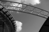 A black and white image featuring a large steel structural arch with visible trusses extending across the sky. Below, there is a section of a modern building with repetitive geometric patterns. Scattered clouds in the sky add texture to the composition.