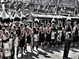 A group of people in traditional attire are gathered outdoors. They wear elaborate headdresses with feathers, beaded jewelry, and intricate clothing. The setting appears to be a cultural event or festival with structures made of natural materials in the background.