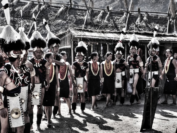 A group of people in traditional attire are gathered outdoors. They wear elaborate headdresses with feathers, beaded jewelry, and intricate clothing. The setting appears to be a cultural event or festival with structures made of natural materials in the background.