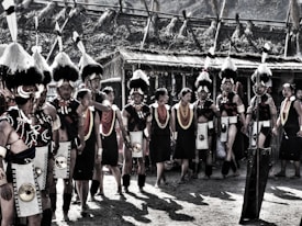 A group of people in traditional attire are gathered outdoors. They wear elaborate headdresses with feathers, beaded jewelry, and intricate clothing. The setting appears to be a cultural event or festival with structures made of natural materials in the background.