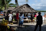 A bustling outdoor market scene by the seaside with vendors selling various fruits under thatched huts. People are interacting and moving around, surrounded by palm trees and the ocean in the background.