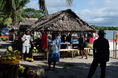 A bustling outdoor market scene by the seaside with vendors selling various fruits under thatched huts. People are interacting and moving around, surrounded by palm trees and the ocean in the background.