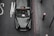 A black taxi is parked on a road next to a curb, viewed from above. The taxi has a classic look with a reflective surface showing minimal reflections of the surroundings. There are visible decals on the windshield and the roof sign reads 'TAXI'. The road has worn white letters and red lines along the curb.