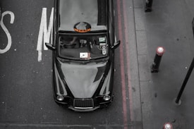A black taxi is parked on a road next to a curb, viewed from above. The taxi has a classic look with a reflective surface showing minimal reflections of the surroundings. There are visible decals on the windshield and the roof sign reads 'TAXI'. The road has worn white letters and red lines along the curb.