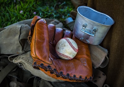 A worn leather baseball glove with a baseball resting inside it. Nearby, a metal camping mug sits partially over a canvas bag. The scene is set in a natural outdoor setting with green grass in the background.